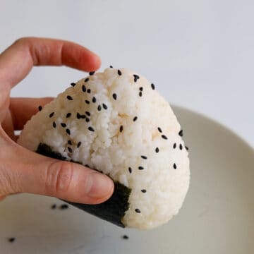 Remove the triangle rice ball from the plastic wrap and stick the rough side of the nori strip on the base of the ball starting from the middle down. Garnish with black sesame seeds and enjoy fresh!