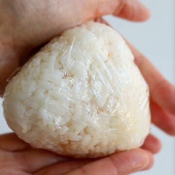 Lift and twist the corners of the plastic wrap so you form a circular rice ball while pushing the filling into the center. Gently flatten the rice ball until it’s about 1.75 inches thick. Then shape it into a triangle using the L-shape of your thumb and pointer finger until you’re satisfied with the shape.