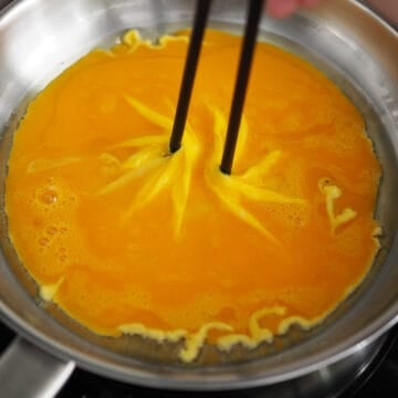 In a small frying pan (about 8 inches wide) set over low-medium heat, add oil. Pour your egg mixture into the pan and let it bubble to create a solid base. Quickly insert wooden chopsticks positioned at a 45-degree angle from each other along the edges of the pan. Then slowly bring chopsticks toward the center.