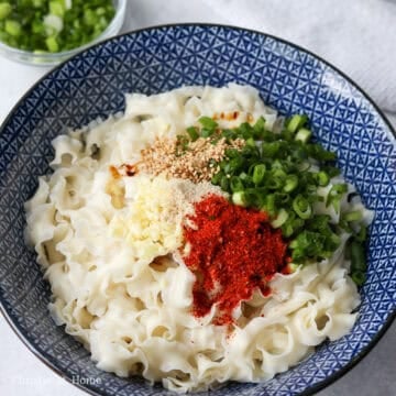 In the centre of the noodles, add regular and dark soy sauce, Chinese black vinegar, garlic, chicken bouillon powder, white granulated sugar, green onions, red chili flakes, sesame seeds and set aside. 